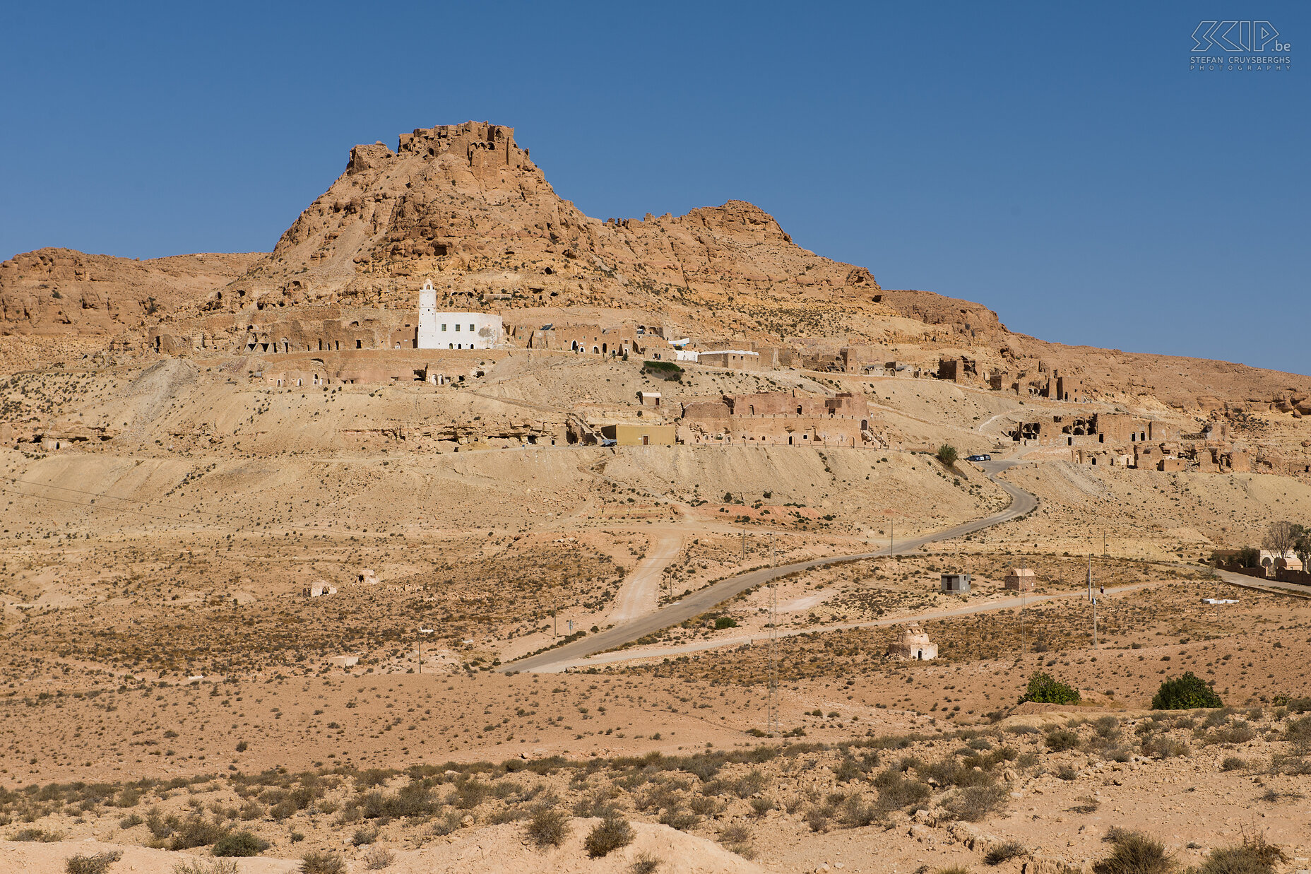 Douiret The abandoned Berber village of Douiret, which has been cut out of the hillside, was once a caravan relay post between Ghabès and Ghadamis. Almost invisible from the ground below the village of Douiret merges into the landscape for hundreds of metres along the mountain. The most impressive buildings of Douiret are since long abandoned. They can rise 3 to 4 storeys high. Some rooms have decorative paintwork and carvings on the roof. Around 1850 about 3,500 people lived there. Today, only 5 families, with 20-30 members keep up the tradition. The main income of Douiret come from olives, and the oil is pressed here by camel-powered constructions. Stefan Cruysberghs